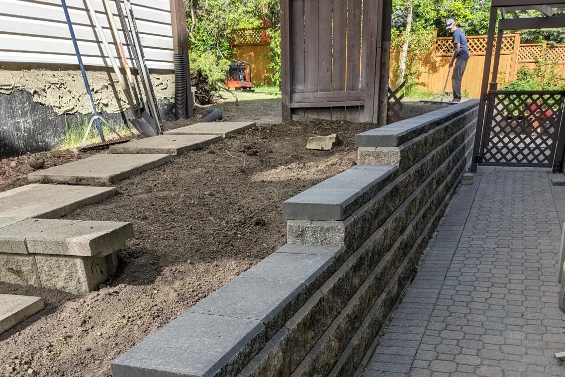 Block retaining wall under construction with crew raking grade, Calgary hardscaping by All The Moore