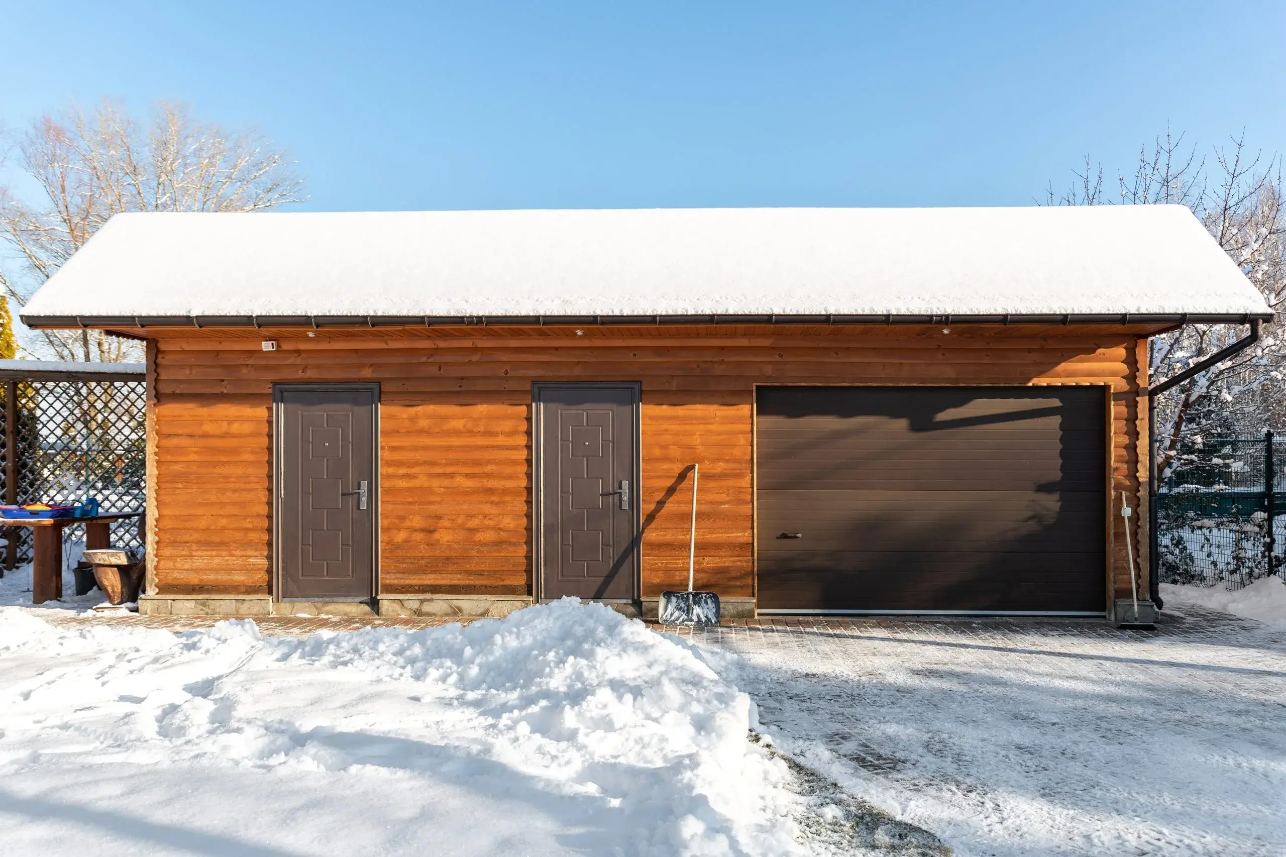 A wooden garage sits covered in fresh snow under a clear blue sky
